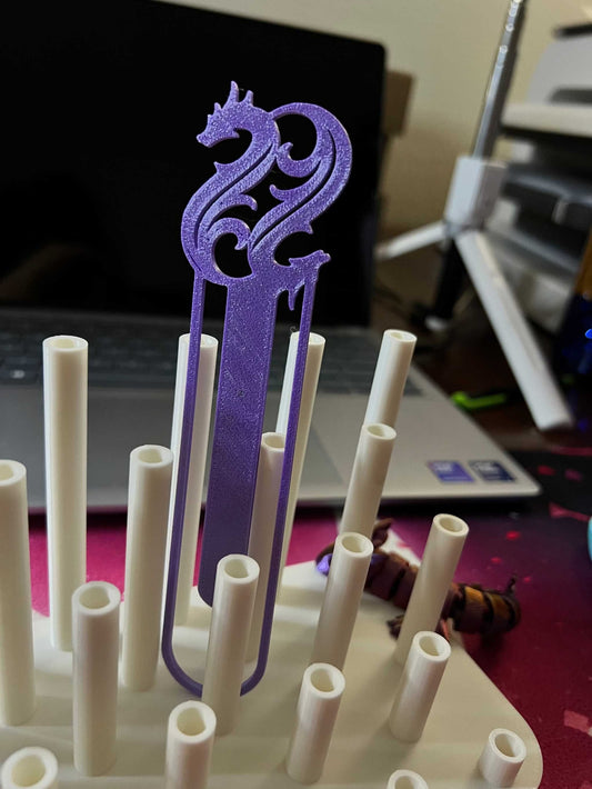 Purple 3D dragon bookmark standing upright on a white pegboard with a laptop in the background