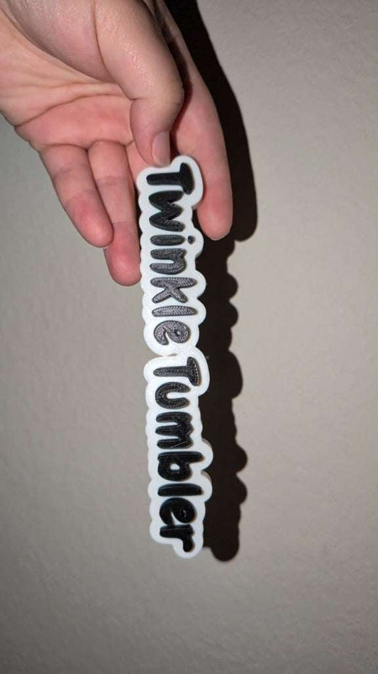 Hand holding a white and black watermark tag name keychain with text "TwinkleTumbler" against a neutral wall background